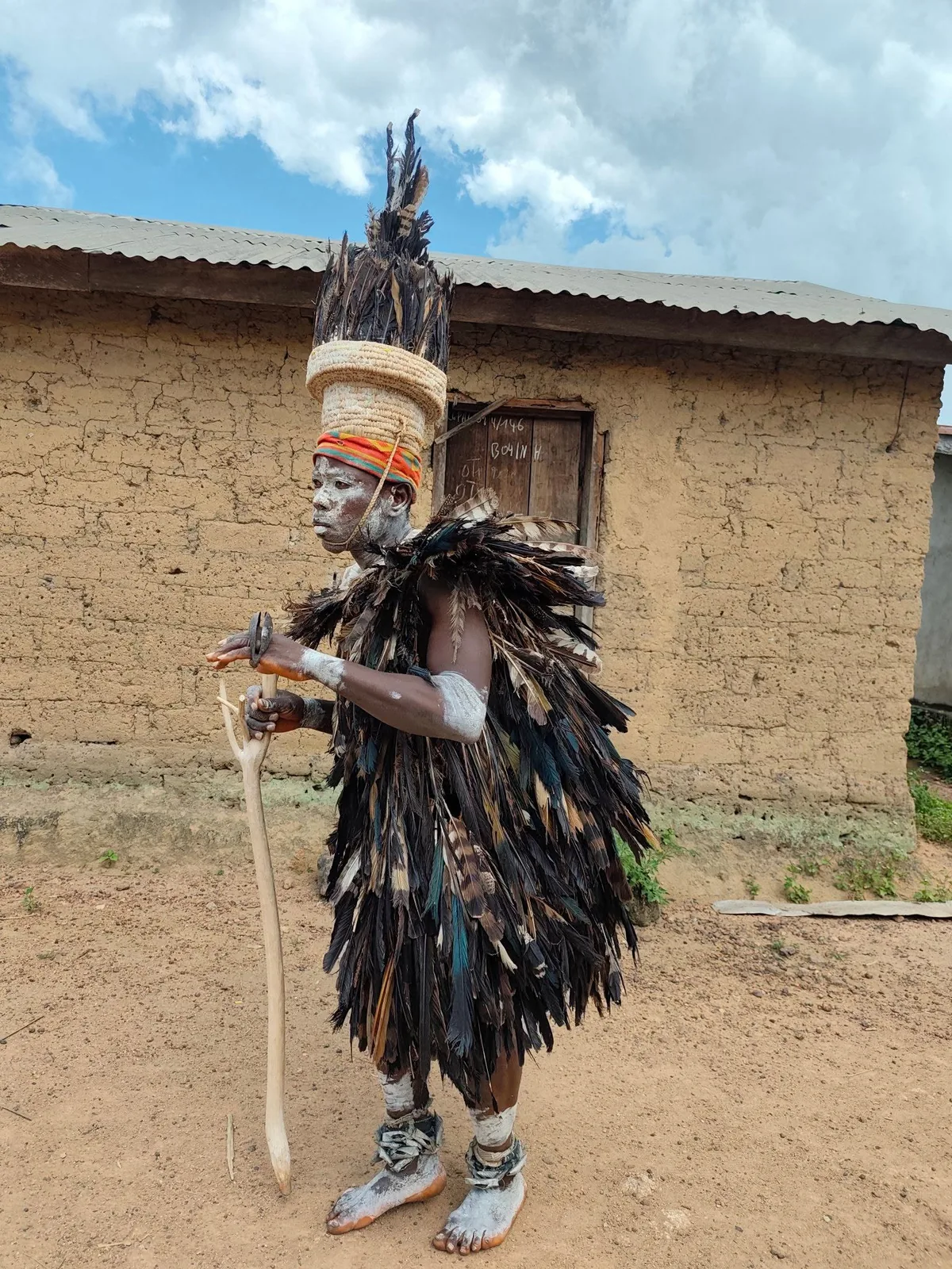 Masked dancer in a full feather costume before a mud-brick house