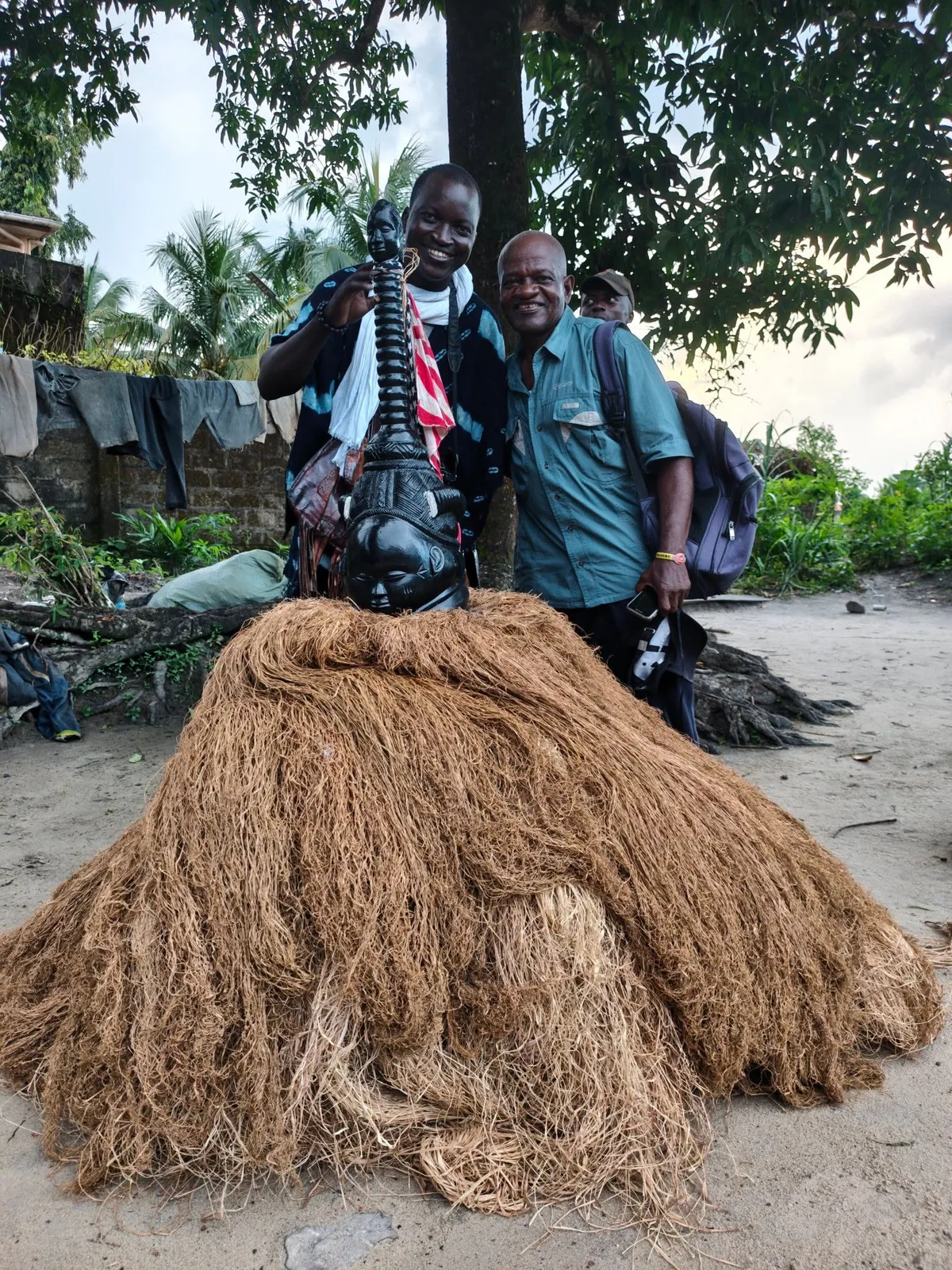 Two men beside a large raffia masquerade costume laid out on the ground